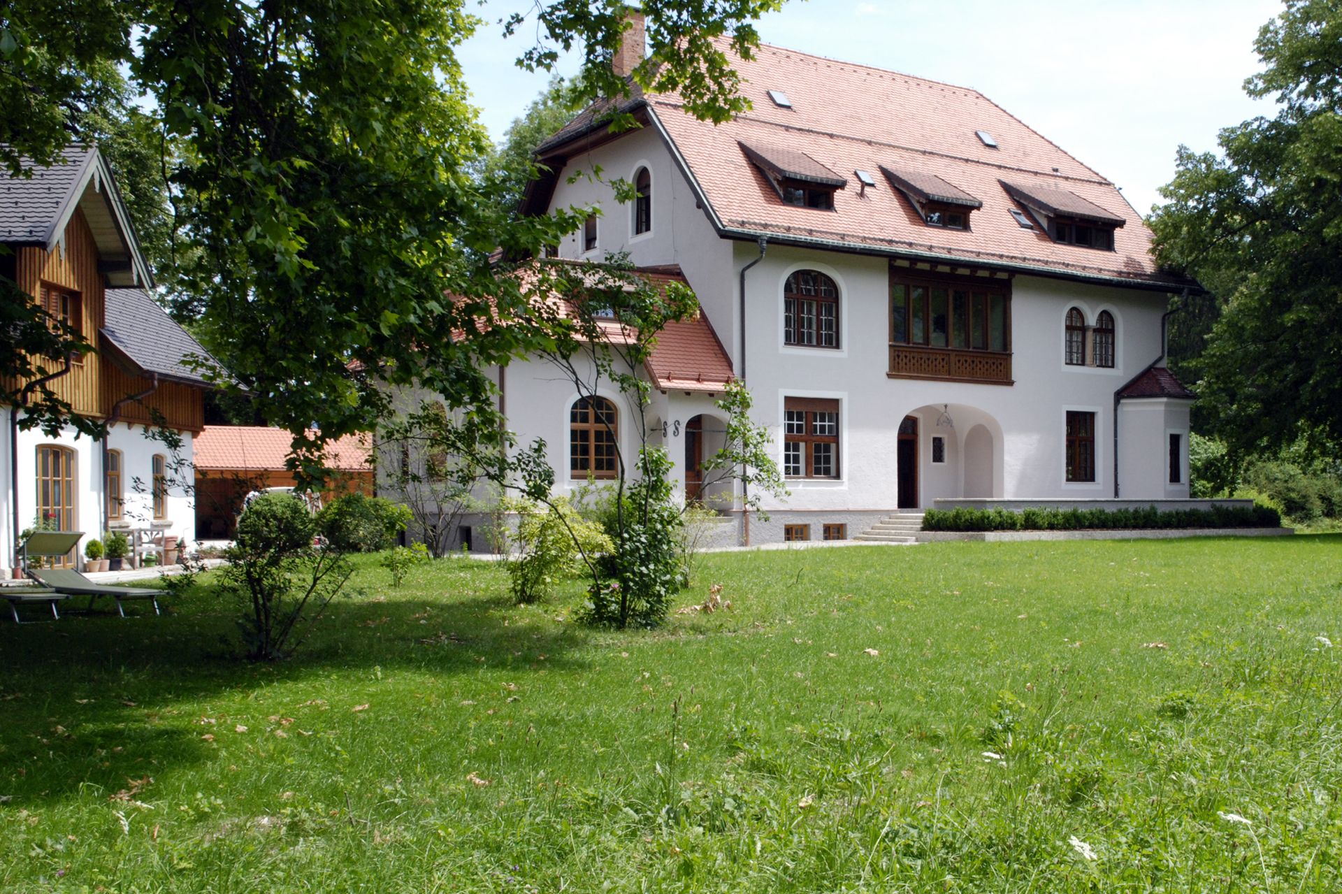3-stöckige Villa mit hellgrauer Außenwand, Holz-Sprossenfenstern, rotem Giebeldach und angrenzender Rasenfläche und Baumbestand