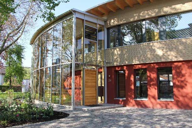 Seitenansicht des Instituts für Baubiologie in Rosenheim mit gelber und roter Fassade sowie großen Glasfronten, das durch energetische Sanierung modernisiert wurde
