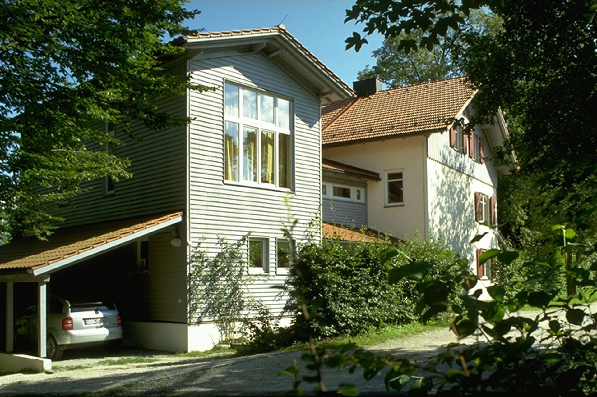 3-stöckiges Wohnhaus, teils in weißer Farbe, teils in grauer Holzlattenbauweise, mit orangem Giebeldach und angrenzendem Carport